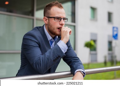 Portrait Of Young Businessman With Beard Standing In Front Of Office Block. He Is Eating Candy Bar. Outdoor Photo.