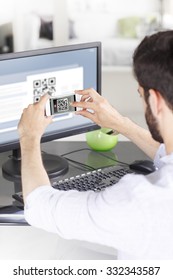 Portrait Of Young Businessman Sitting At His Workplace In Front Of Computer Monitor And Scanning QR Code With Mobile Smart Phone. 