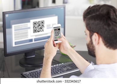 Portrait Of Young Businessman Sitting At His Workplace In Front Of Computer Monitor And Scanning QR Code With Mobile Smart Phone. 