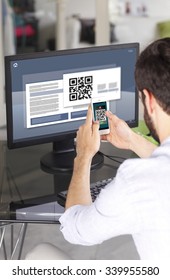 Portrait Of Young Businessman Sitting At His Workplace In Front Of Computer Monitor And Scanning QR Code With Mobile Smart Phone. 