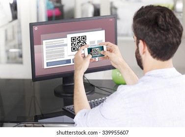 Portrait Of Young Businessman Sitting At His Workplace In Front Of Computer Monitor And Scanning QR Code With Mobile Smart Phone. 