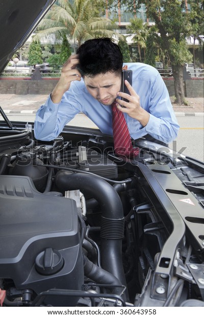 Portrait of young businessman talking on the\
phone while checking the broken machine\
car