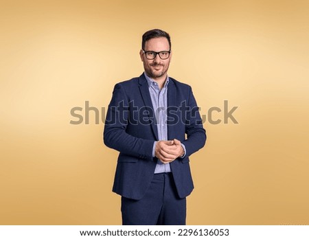 Portrait of young businessman wearing glasses and elegant blue suit clasping his hands. Male professional posing confidently while standing isolated on beige background
