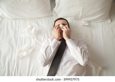 Portrait Of Young Businessman Wearing White Shirt And Necktie Lying On The Bed In The Hotel Room Sick With Flu. Caught Cold And Sneezing Into Tissue. View From Above