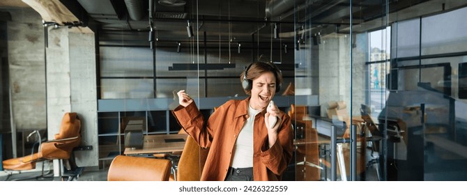 Portrait of young businesswoman, employer singing at smartphone in her office, listening music in wireless headphones, having fun at her workplace. - Powered by Shutterstock