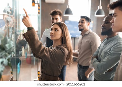 Portrait of young businesswoman mentor coach leader writing idea or task pointing at sticky notes on glass wall, diverse team developing work plan in creative corporate office at stand up meeting