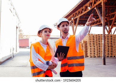 Portrait Of Young Businesswoman Near Professional Engineer Both In Orange Vests And Hard Hats Outdoor At The Woodworking Factory. Young Engineer Explaining And Showing Something To Female Boss.