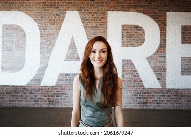 Portrait of a young businesswoman standing in front of an office brick wall with the word dare written on it