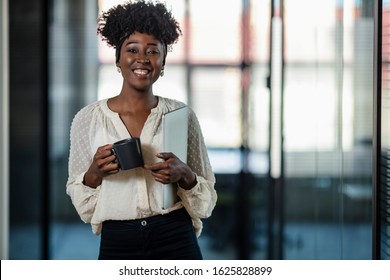 Portrait Of Young Businesswoman Standing With Her Coffee And Laptop In An Office. Portrait Of A Young African Ethnicity Businesswoman. Woman With Friendly Personality, Smiling Isolated Indoors Office