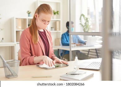 Portrait Of Young Businesswoman Wearing Mask Sanitizing Laptop While Working At Desk In Cubicle At Post Pandemic Office, Copy Space