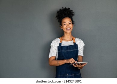 Portrait of young cafe black waitress using digital tablet on grey background. Happy young black woman working on digital tablet while looking at camera. African american cafe owner on gray wall. - Shutterstock ID 2143243891