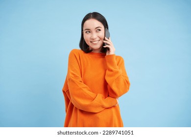 Portrait of young candid woman, asian girl talking on cellphone, holds mobile phone and speaks, stands over blue background. - Powered by Shutterstock