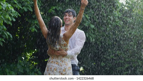 Portrait of young carefree couple in love are dancing and kissing under the rain on a background of green trees.