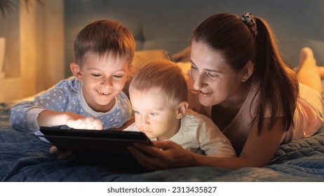 Portrait of young caring mother with two sons using tablet computer in bed before going to sleep. Family having time together, parenting, happy childhood and entertainment - Powered by Shutterstock