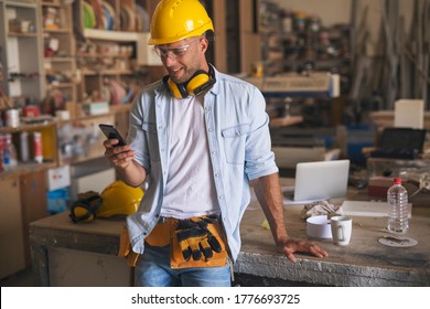 Portrait of a young carpenter wearing helmet, ear protection, protective goggles and a tool belt. He is at his workplace, holding cellphone, texting and smiling.