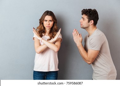 Portrait Of A Young Casual Couple Standing Together Isolated Over Gray Wall Background, Man Asking For Something And Woman Refuses