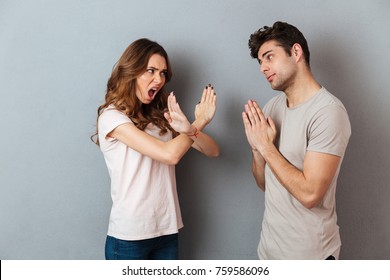 Portrait Of A Young Casual Couple Standing Together Isolated Over Gray Wall Background, Man Asking For Something And Woman Refuses