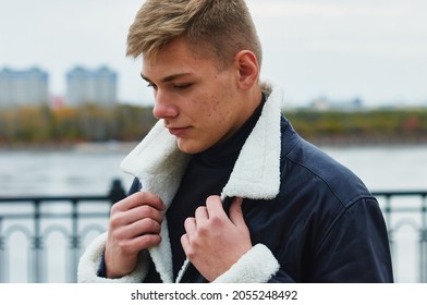 Portrait Of A Young Caucasian Blond Guy In A Warm Leather Black Jacket. The Guy Lifts The Fur Collar With Both Hands. Skin Acne.