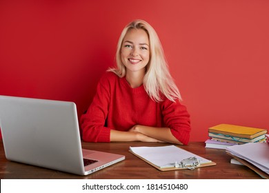 Portrait Of Young Caucasian Blonde Female At Work Place, She Sits In Casual Wear At Office Desk, Enjoy Working On Laptop And With Documents. Isolated Red Background