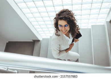 Portrait of young Caucasian businesswoman wearing high-heeled shoes walking upstairs and using digital tablet. Business concept. Cute young businesswoman walking up stairs