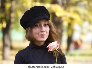 Portrait of an young caucasian cheerful smiling woman dressed in a black peak cap looking at you and holding a flower in her hands. Autumn park and yellow foliage serves as a blurred background.