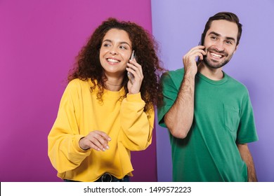 Portrait of young caucasian couple man and woman in colorful clothing smiling and talking on cellphones isolated over violet background