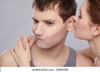 Portrait Of Young Caucasian Couple On Gray. Portrait Of Young Caucasian Couple On Gray. Girl Is Telling Something On Man's Ear And He Is Looking Dissapointed