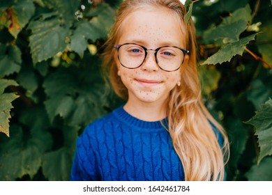 Portrait of young caucasian handsome girl with long fair hair in blue hoody with glasses stands in the garden on the background of green leaves - Shutterstock ID 1642461784