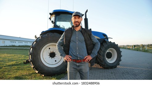 Portrait Of Young Caucasian Handsome Man Farmer In Cap Standing On The Road In Field And Smiling To Camera. Big Tractor On Background. Male Worker In Agricultural Farm. Agriculture Farming.