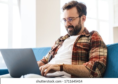 Portrait of young caucasian man wearing plaid shirt using laptop while sitting on sofa at home