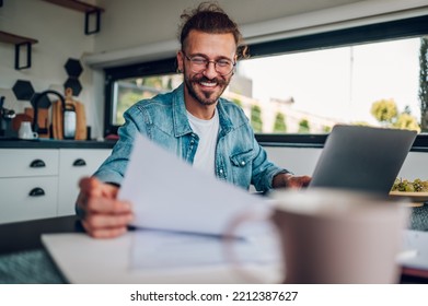 Portrait Of A Young Caucasian Tattooed Man Sitting At The Table In The Kitchen At Home And Working. Using Laptop And Holding Work Documents. Bearded Male Wearing Eyeglasses And Jeans Shirt.