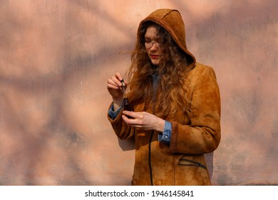 A portrait of a young caucasian woman with light brown blond curly hair holding an open CBD oil bottle with a dipper in her hand. The girl is wearing a red lipstick, a jeans shirt and a brown jacket.