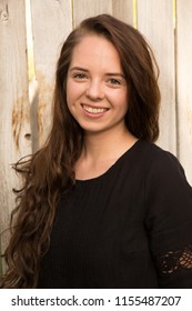 Portrait Of A Young Caucasian Woman With Long Brown Hair Wearing A Black Dress Standing Next To A Rustic Old Wooden Fence.  Model Head Shot Of Young Woman, Brunette Hair. Soft Natural Evening Light.