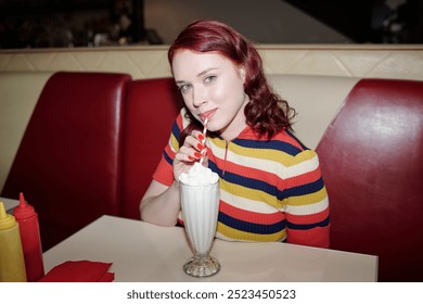 Portrait of young Caucasian woman sipping milkshake while sitting in retro diner booth, with red and beige upholstery and condiments on table - Powered by Shutterstock