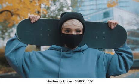 Portrait of young caucasian woman skater with protective face mask holding skateboard behind her head in the park in autumn. High quality photo - Powered by Shutterstock