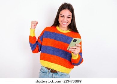 Portrait of Young caucasian woman wearing colorful sweater over white background holding in hands cell celebrating