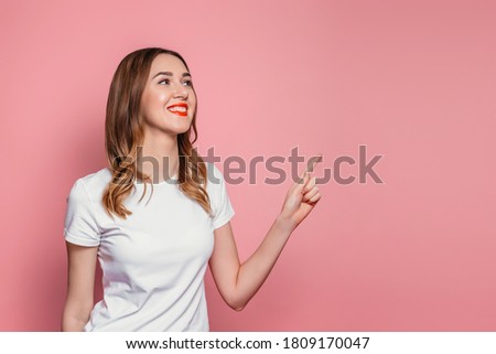 Portrait of young caucasian woman in white t-shirt with red lipstick pointing with finger at copy space for text, girl looking at background