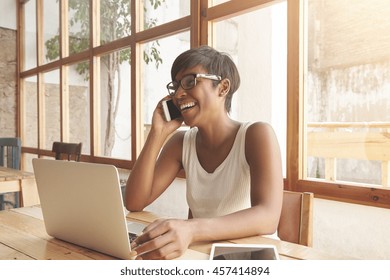 Portrait Of Young Charismatic African Adult Woman Laughing While Talking On Cellphone With Her Man, Remembering Good Old Days And Their First Date, Black Woman Sitting At Cafeteria Using Laptop