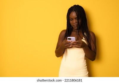 Portrait Of Young Cheerful African Woman Using Smartphone For Messaging With Friends, Happy African Lady Texting On Cellphone While Standing Over Yellow Studio Background, Copy Space