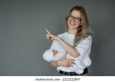 Portrait Of Young Cheerful Attractive Woman Holding Baby Doll In White Clothes, Pointing With Index Finger On Grey Background. Motherhood, Childbirth, Breastfeeding, Breast Model, Fake, Education. 