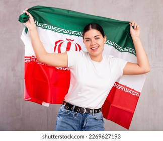 Portrait of young cheerful female in casual wear holding national flag of Iran against gray wall