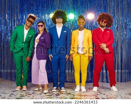 Portrait of young cheerful friends posing at theme party against shiny blue foil curtain. Multiracial people in colorful clothes, sunglasses and wigs stand in row and smile at camera. Full length.