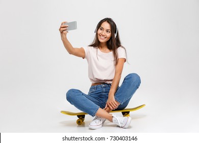 Portrait of young cheerful girl in white headphone makes selfie on smartphone, sitting on yellow skateboard, isolated over white background - Powered by Shutterstock