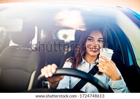 Portrait of a young and cheerful woman with coffee cup driving luxury car in the city. Smiling pretty young woman drinking take out coffee when driving to work in the morning