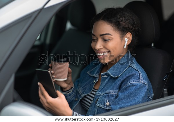 Portrait of young and cheerful woman with coffee
cup sits in car in city on break. Smiling pretty african american
lady in wireless headphones, look at phone, drinking takeaway
coffee in morning
