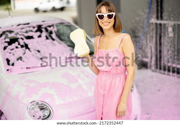 Portrait of a young cheerful woman dressed in\
pink dress standing with sponge in front of her tiny car in foam at\
self-service station