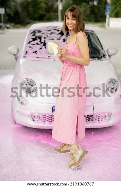 Portrait of a young cheerful woman dressed in pink\
dress standing in front of her tiny car in pink foam at\
self-service car wash