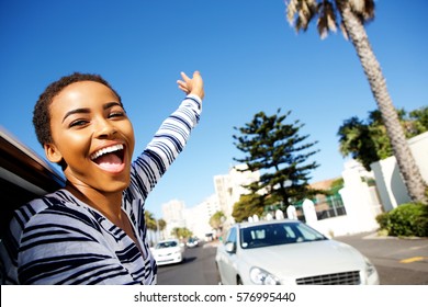Portrait of young cheerful woman hanging outside car window with arms raised