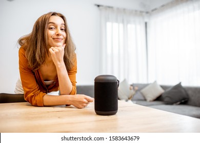 Portrait Of A Young And Cheerful Woman With A Smart Wireless Column At Home. Concept Of Smart Home And Voice Command Control