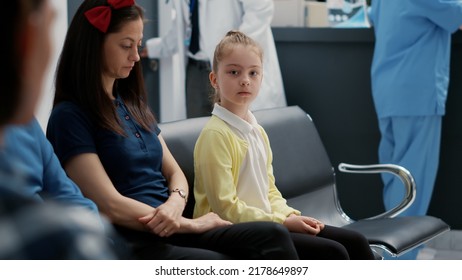 Portrait Of Young Child Sitting With Mother In Waiting Area At Hospital Reception, Attending Annual Checkup Visit With Specialist. Little Girl In Waiting Room Lobby At Healthcare Facility.